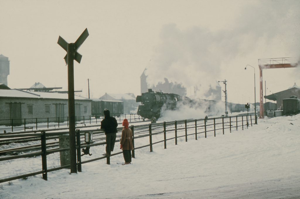 Bad Oldesloe: Bahnhof: Gleisanlagen: davor Geländer mit zwei Kindern: links Andreaskreuz: in der Mitte Dampflokomotive mit Güterzug: hinten links Schuppen: dahinter weitere Gebäude und Wasserturm: hinten rechts Güterbahnhof: im Hintergrund Mitte Silos der Firma Karl Klüßmann und der Johannes Ströh Mühlenwerke: Winter, 1960