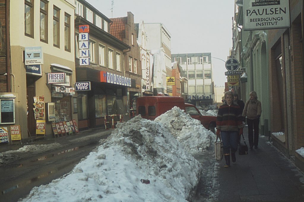 Bad Oldesloe: Hindenburgstraße: vorn Schneehaufen und parkender Pkw: rechts Fußgänger vor Beerdigungsinstitut Paulsen, Apotheke, Taxiunternehmen: links weitere Wohn- und Geschäftshäuser mit Friseursalon, Zeitschriftenladen, Volksbank Bad Oldesloe, Schuhgeschäft, Commerzbank: hinten Kaufhaus Nickel an Mühlenstraße: Winter, 1979